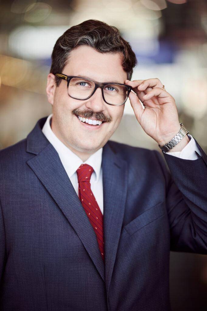 Porträtfoto eines Mannes in Anzug, der mit seiner Hand die Brille berührt.